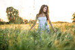 © Designpics - Young girl stands in the rye field and looks to the left on the sunset background. Woman touches the rye. She wears a light dress with prints of flowers. Outdoors. Horizontal.