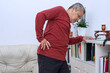 Elderly male grandpa near sofa at home holding hands on back having painful muscle and lower backache injury problem.