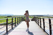 © @skuder_photographer - Beautiful young woman with long brown hair dressed in short skirt and shirt crosses the wooden bridge doing different poses like a model. In the background the blue sky and white clouds. She is happy.