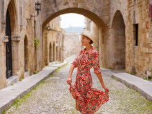 Summer Trip To Rhodes Island, Greece. Young Asian Woman In Ethnic Red Dress Walks Street Of Knights Of Fortifications Castle. Female Traveler Visiting Southern Europe. Unesco World Heritage Site.