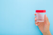 Young adult woman hand holding transparent plastic container for urine or other analysis test on light blue background. Pastel color. Healthcare concept. Closeup. Empty place for text. Front view.