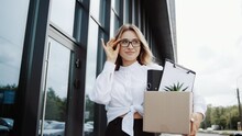 Business woman in glasses walking outdoor after unemployment from company. Attractive girl employee worker hold a box, feel sad and upset after losing the job due to world economic problem