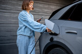 Fototapeta Sypialnia - Young woman plugs charging gun in her car while standing with a smart phone near garage doors of her house. Concept of green and cheap energy of domestic vehicle charging