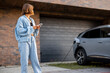 Young woman useing smart phone while charging her electric car near garage of her house. Concept of technologies and EV cars