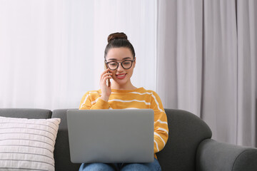 Wall Mural - Home workplace. Happy woman with laptop talking on smartphone on sofa in room