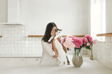 Happy woman hugging cute cat at peonies in vase on background of minimal white kitchen in new modern home. Housewife decorating house with flowers and playing with cat, housekeeping