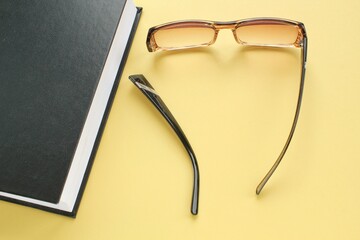 Old broken eyeglasses with damaged legs and books on yellow background. Poor eyesight. Repair concept. Idea of health. Failure optic eyewear. Breakage of vision correction glasses. Close up, flat lay