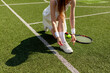young girl tennis player in white sports uniform ties her shoelaces on sneakers on green court, woman coach