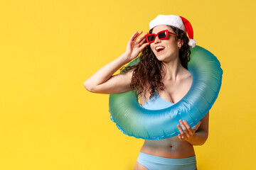 Wall Mural - attractive girl in swimsuit and sunglasses in santa hat and with an inflatable swim ring smiles on yellow background