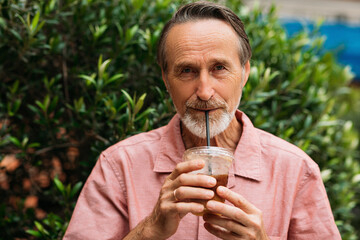 Sticker - Senior man drinking a smoothie and looking at camera while standing on the park