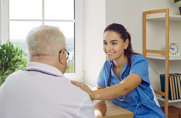 Wall Mural - Happy friendly young doctor, caregiver or nurse in blue uniform scrubs smiling and touching senior man's shoulder. Medicine, senior care, retirement home, medical help, moral support concept