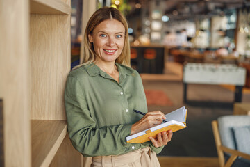 Wall Mural - Pretty woman entrepreneur making notes in note pad while standing on cozy coworking background