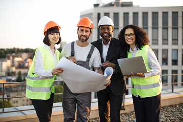 portrait of multicultural group of developers posing on camera with portable gadget and blueprints. 