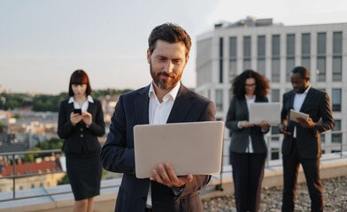 Handsome investor in business wear with laptop looking straight while standing on office terrace with fantastic city view. International team of supervisors using gadgets for designing new project.