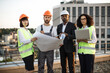 © sofiko14 - Portrait of multicultural group of developers posing on camera with portable gadget and blueprints. Four international colleagues in vests and hardhats smiling while meeting at panoramic terrace.