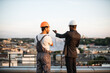 © sofiko14 - Back view of foreman in grey uniform and business investor in black suit watching together at engineering drawings while standing on panoramic terrace. Male builders discussing real estate project.