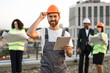 Portrait of cheerful male constructor with digital tablet in hand touching hardhat and smiling at camera. Multiracial team of urban planners watching at paper design layout on blurred background.