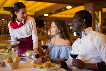 Wall Mural - Smiling waitress serving food for positive couple at restaurant, putting plate with salad on table