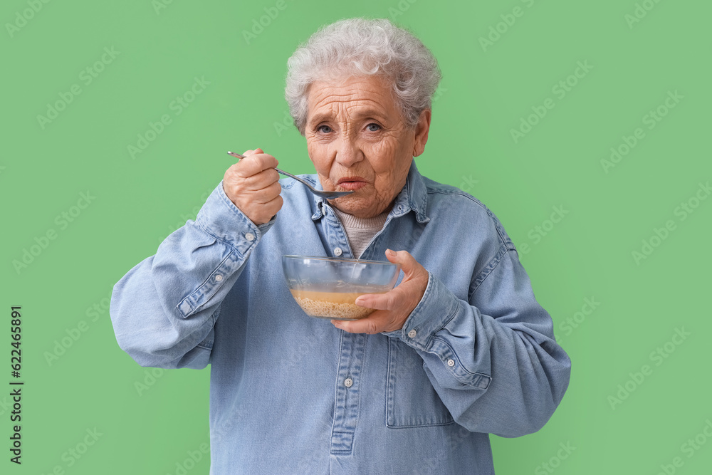 Ill senior woman eating soup on green background
