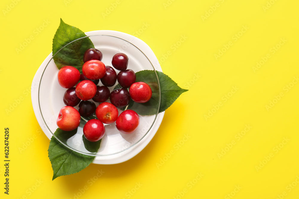 Plate with sweet cherries and leaves on yellow background