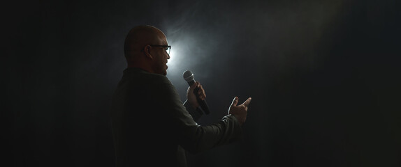 Canvas Print - Confident successful speaker man talking on stage with spotlight strike through the darkness at corporate business event. Public speaker giving talk at conference hall. Stand up comedian.