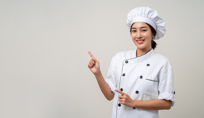 Wall Mural - Smiling Young beautiful asian woman chef in uniform pointing finger to copy space isolated background. Cooking woman Occupation chef or baker People in kitchen restaurant and hotel.