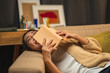 © Chanakon - Asian beautiful woman lay down on cozey couch sofa reading book in living room with warm light at night. People female stay home part time relax at home.