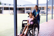 Happy diverse schoolgirl in wheelchair with her friend in corridor at school