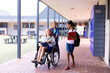 Happy diverse schoolgirl in wheelchair with her friends in corridor at school
