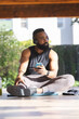 © Wavebreak Media - Thoughtful african american man wearing sportswear, headphones and using smartphone on mat in garden