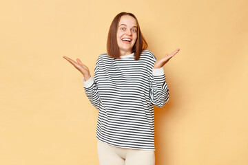 Wall Mural - Surprised happy brown haired woman wearing striped shirt standing isolated over beige background posing with raised arms looking with positive amazed expression.