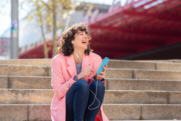 Wall Mural - Cheerful young woman sitting on city steps enjoying listening to music