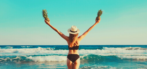 Summer vacation, rear view of beautiful happy slim woman in bikini swimsuit and straw hat with pineapples fruits raising her hands up on the beach on sunny day on sea background