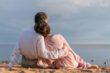 Wall Mural - young woman in pink dress and an elderly man sit hugging together on beach at sunset. concept of the relationship of different ages in a couple