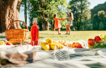 Picnic blanket with boiled corn, fruits, a bottle of juice, basket still life in city summer park with two sisters girls rotating on background holding hand in hand. Family values and outdoors concept
