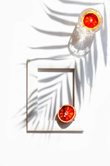 Wall Mural - Creative composition made of half bloody red orange and a glass with cocktail on white background with frame and palm tree leaf shadow. Summer refreshment and party concept. Minimal style. Top view