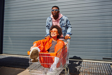 Excited young african american man in sunglasses and denim jacket standing near smiling best friend sitting in shopping cart and building on urban street, friends hanging out together