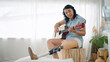 Happy asian woman in leg prosthesis equipment enjoy playing guitar and singing while sitting on chair in living room at home. Leg prosthetic equipment, Amputee concept
