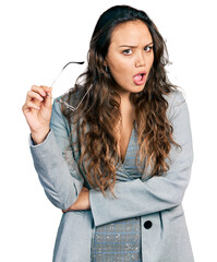 Canvas Print - Young hispanic girl wearing business clothes holding glasses in shock face, looking skeptical and sarcastic, surprised with open mouth