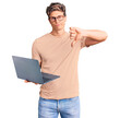© Krakenimages.com - Young handsome man wearing glasses holding laptop with angry face, negative sign showing dislike with thumbs down, rejection concept