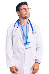 Wall Mural - Young hispanic man wearing doctor uniform and stethoscope smiling looking to the side and staring away thinking.