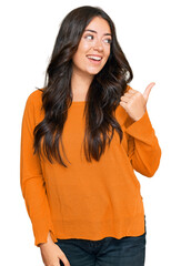 Wall Mural - Beautiful brunette young woman wearing casual orange sweater smiling with happy face looking and pointing to the side with thumb up.