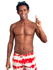 Canvas Print - African handsome man wearing swimsuit and sunglasses showing and pointing up with finger number one while smiling confident and happy.