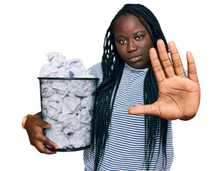 Poster - Young black woman with braids holding paper bin full of crumpled papers with open hand doing stop sign with serious and confident expression, defense gesture