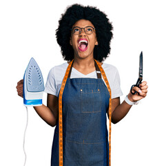 Poster - Young african american woman dressmaker designer wearing atelier apron holding iron and scissors angry and mad screaming frustrated and furious, shouting with anger looking up.