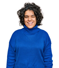 Young hispanic woman with curly hair wearing turtleneck sweater winking looking at the camera with sexy expression, cheerful and happy face.