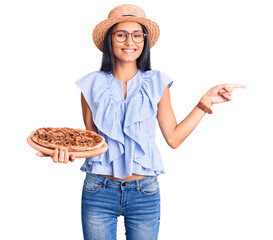 Canvas Print - Young beautiful latin girl wearing summer hat and glasses holding pizza smiling happy pointing with hand and finger to the side