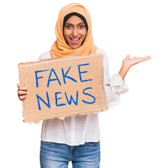 Poster - Young brunette arab woman holding fake news banner celebrating victory with happy smile and winner expression with raised hands