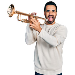 Wall Mural - Young hispanic man with beard playing trumpet sticking tongue out happy with funny expression.