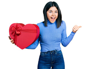 Poster - Young brunette woman with blue eyes holding valentine gift pointing thumb up to the side smiling happy with open mouth